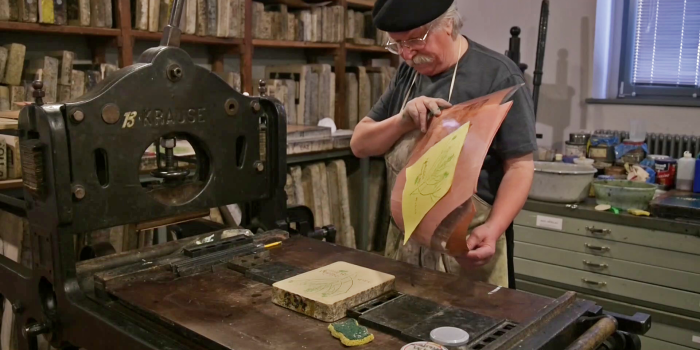 Ein älterer Mann steht vor einer Druckerpresse und hält Papier in der Hand. Auf dem Tisch vor ihm liegt ein Stein.